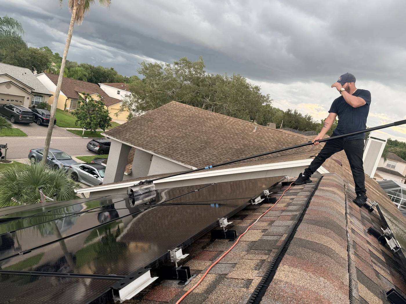 Technician on a shingle roof cleaning a solar array with a long pole brush