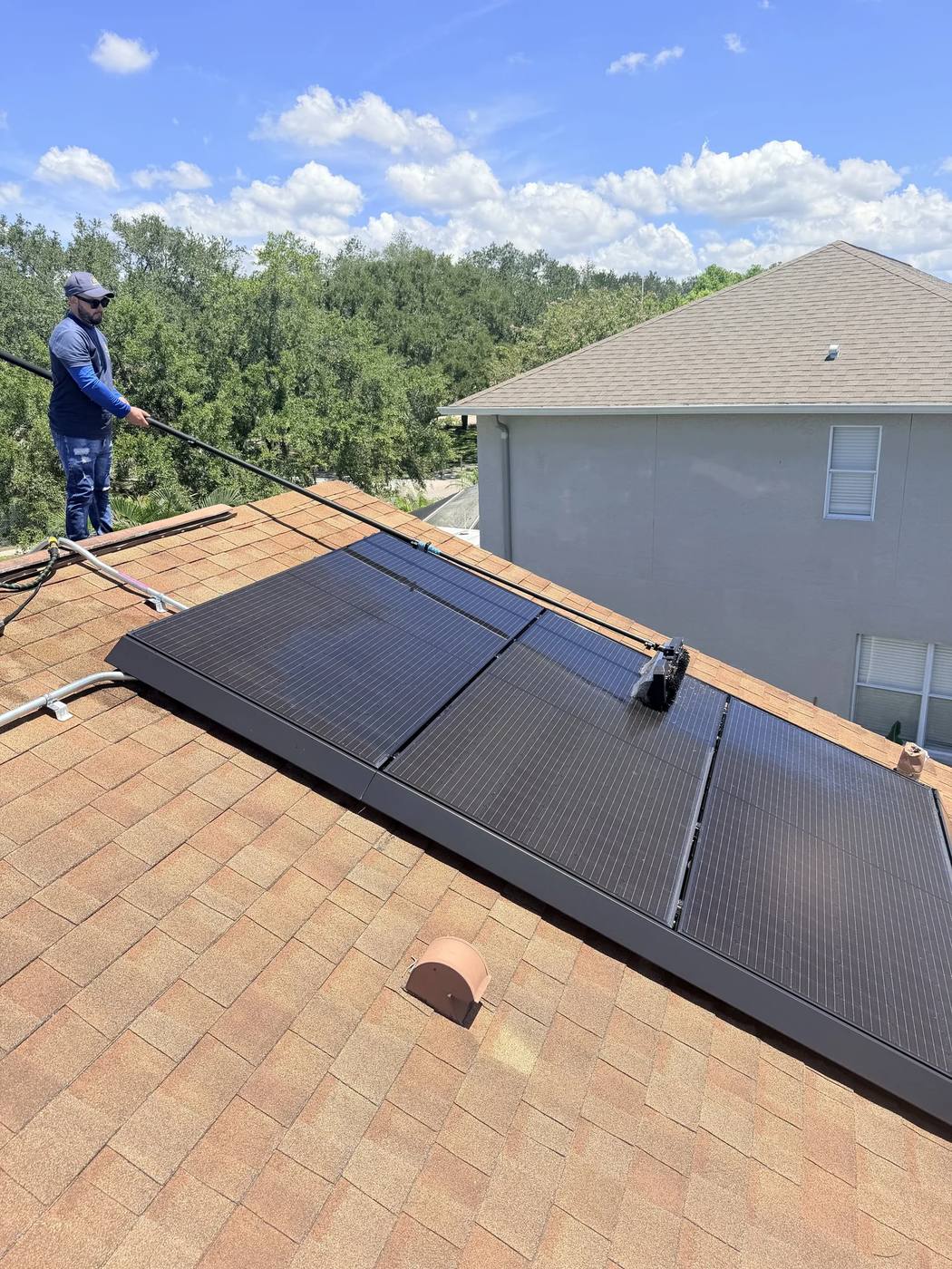 Technician working a pole brush across black solar panels on a shingle roof